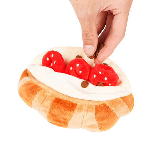Plush toy resembling a croissant with strawberries and cream, held by a hand on a white background.
